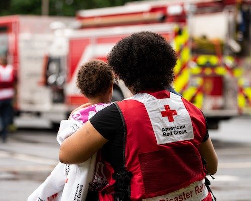 Red Cross volunteer wearing a vest stands overlooking a fire truck while holding an infant wrapped in a blanket
